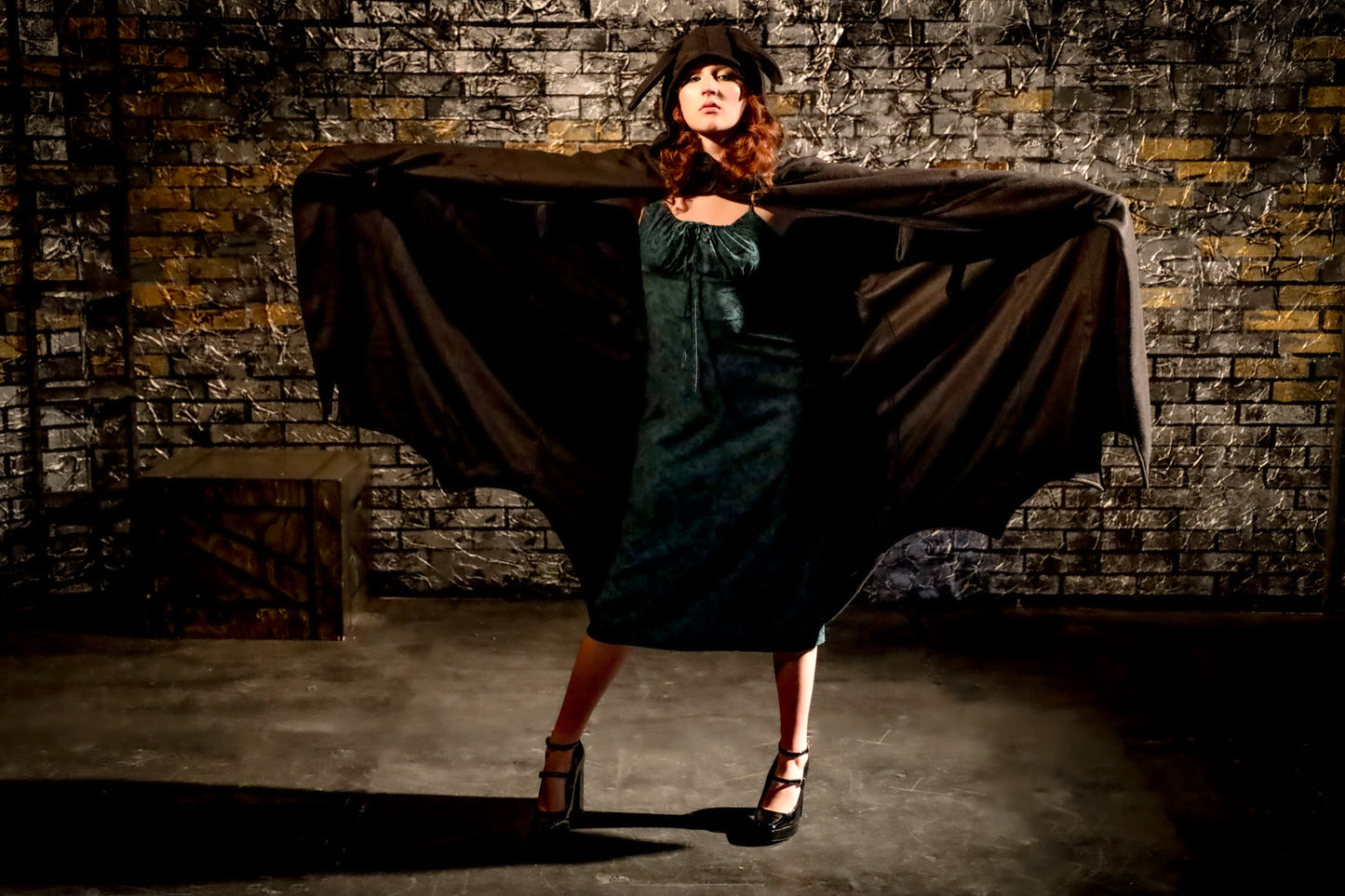 Woman in a dark green dress standing in front of a draped cloth against a brick wall.