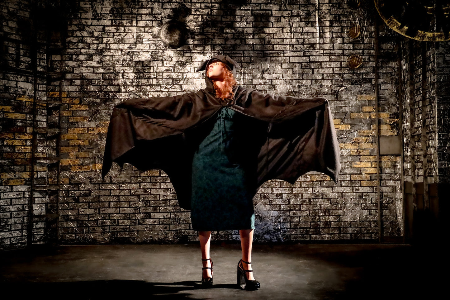 Person in a dark cape and dress standing against a textured brick wall.