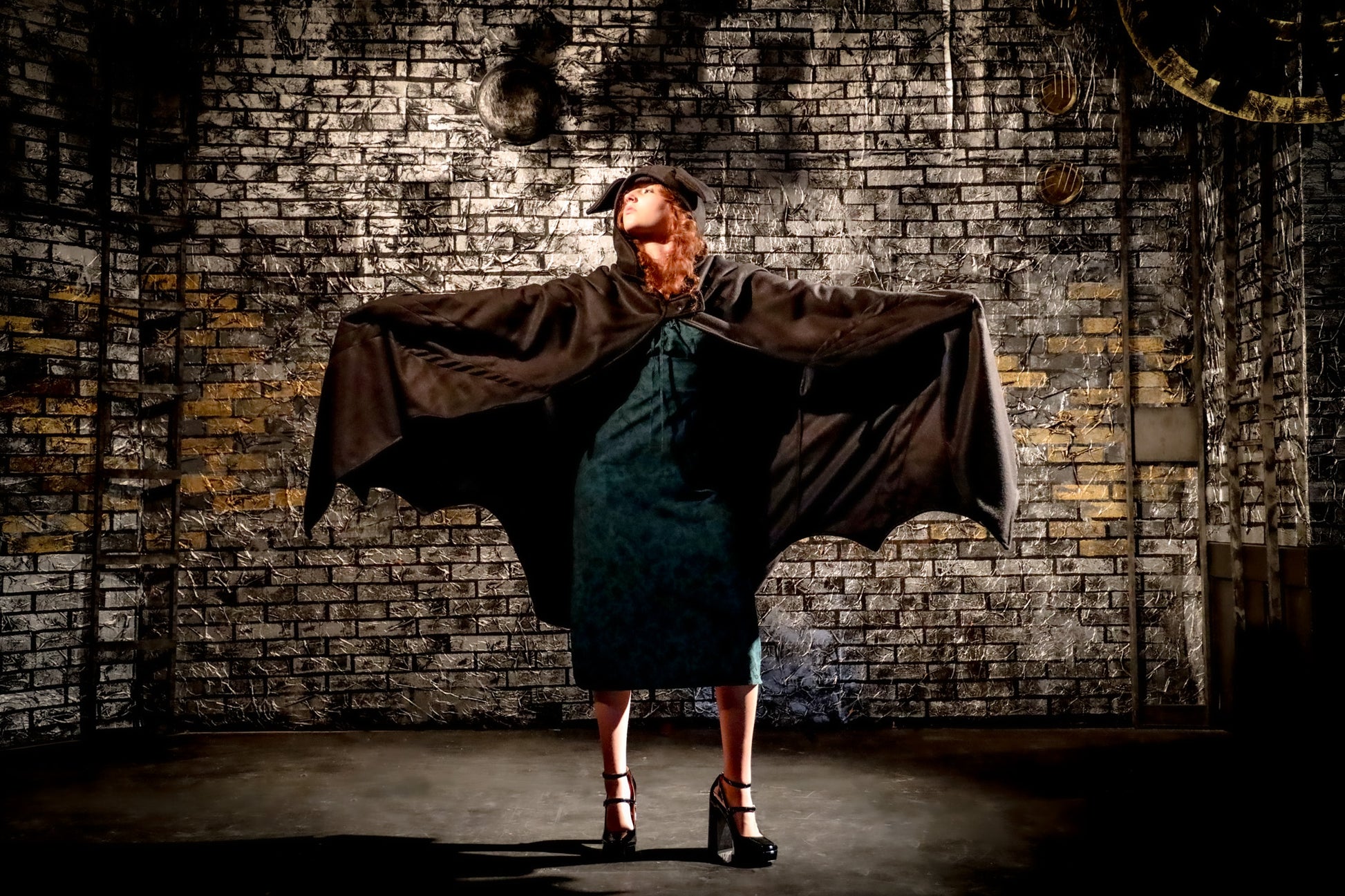 Person in a dark cape and dress standing against a textured brick wall.