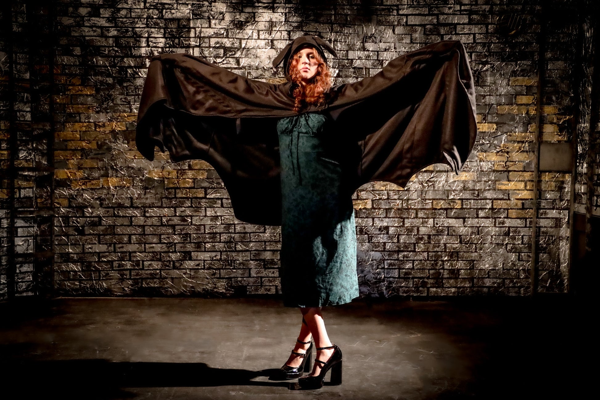 Woman in a dark dress with a cape against a brick wall.