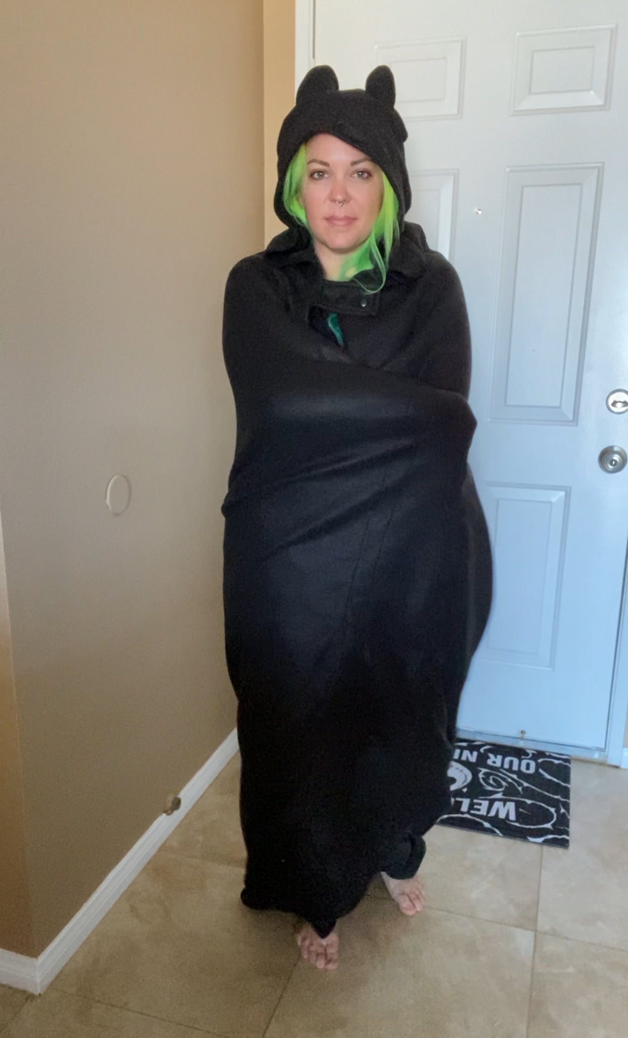 Person wrapped in a black blanket with bat ears in a room.